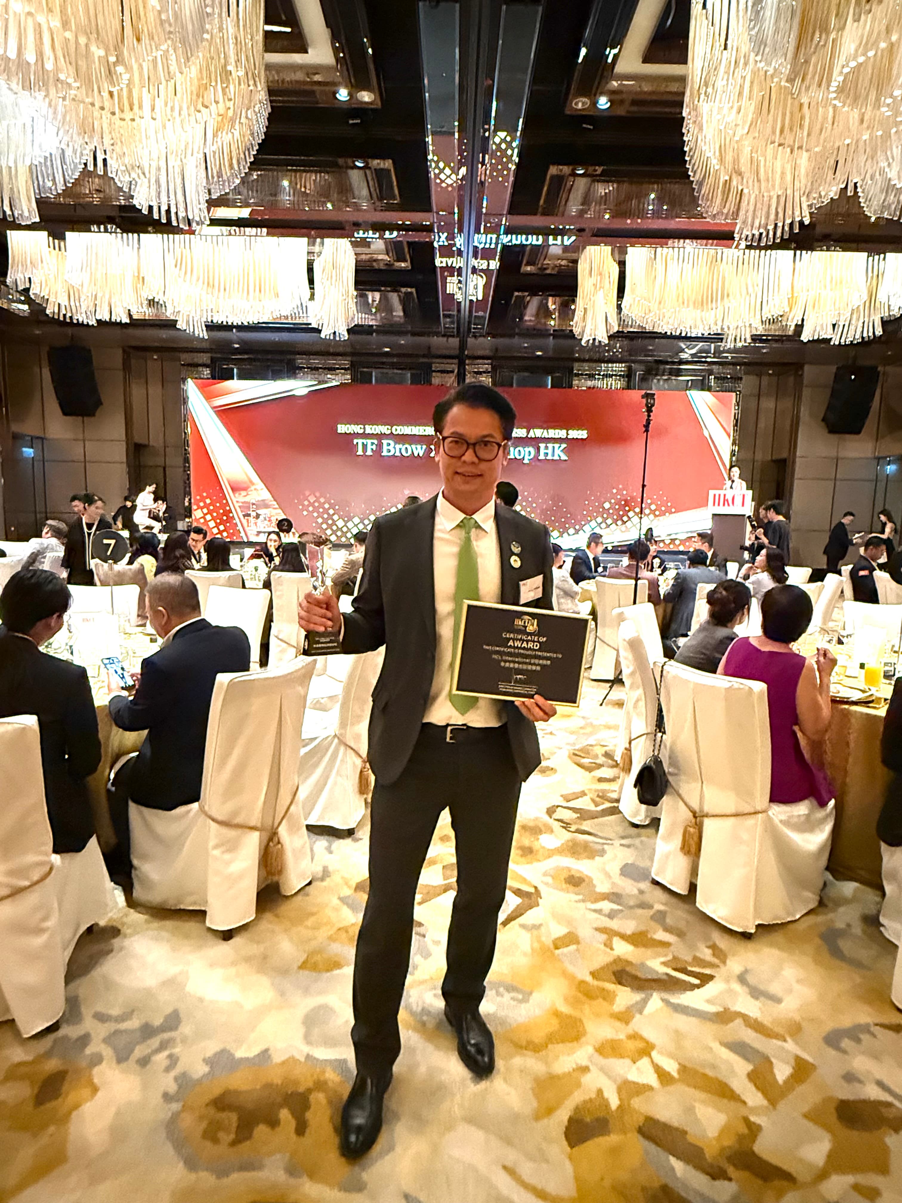 一名穿西装的男子在华丽宴会厅手持奖杯和证书,背景为颁奖典礼舞台。